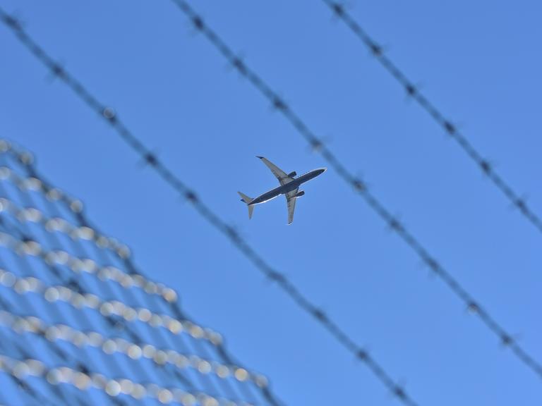 Flugzeug am blauen Himmel, im Vordergrund: ein Zaun mit Stacheldraht