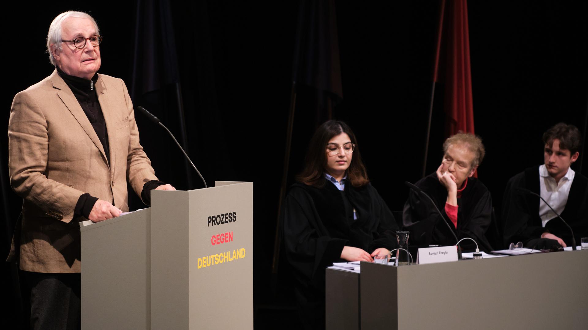 In einer Szene des Stücks "Prozess gegen Deutschland" steht ein Mann an einem Redepult, drei weitere Personen sitzen daneben und hören zu.