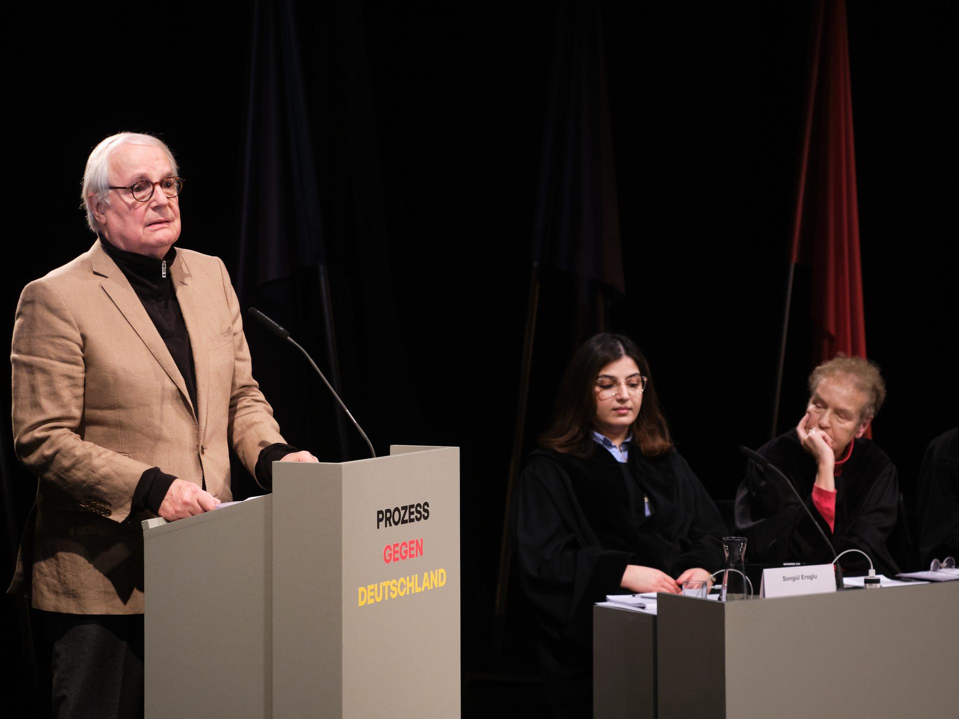 In einer Szene des Stücks "Prozess gegen Deutschland" steht ein Mann an einem Redepult, drei weitere Personen sitzen daneben und hören zu.