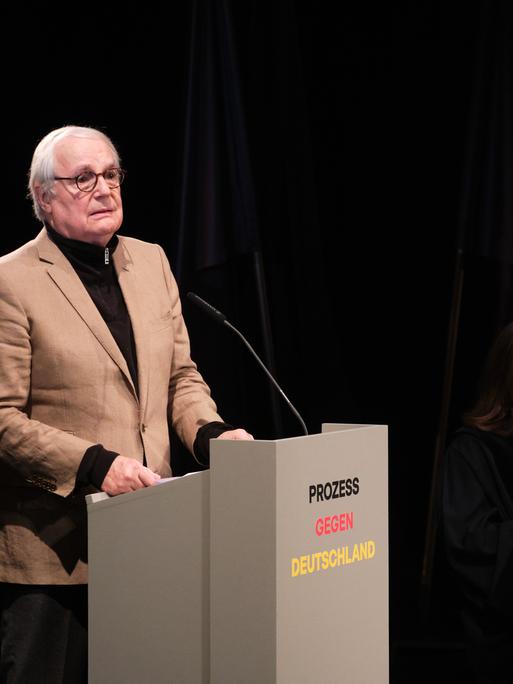 In einer Szene des Stücks "Prozess gegen Deutschland" steht ein Mann an einem Redepult, drei weitere Personen sitzen daneben und hören zu.