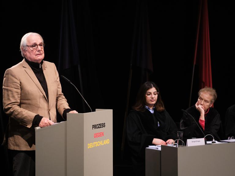 In einer Szene des Stücks "Prozess gegen Deutschland" steht ein Mann an einem Redepult, drei weitere Personen sitzen daneben und hören zu.