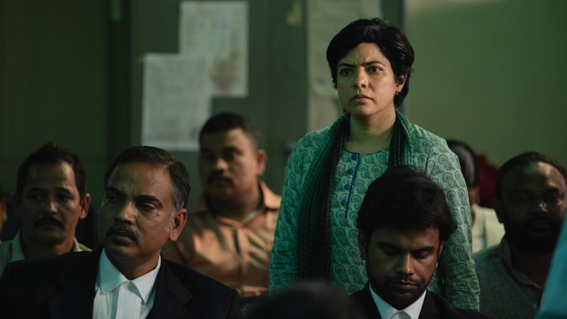 Filmszene aus der Serie "Trial by Fire". Zu sehen ist eine stehende Frau, vermutlich die Hauptdarstellerin, zwischen sitzenden Männern. Vermutlich in einem Gerichtssaal.