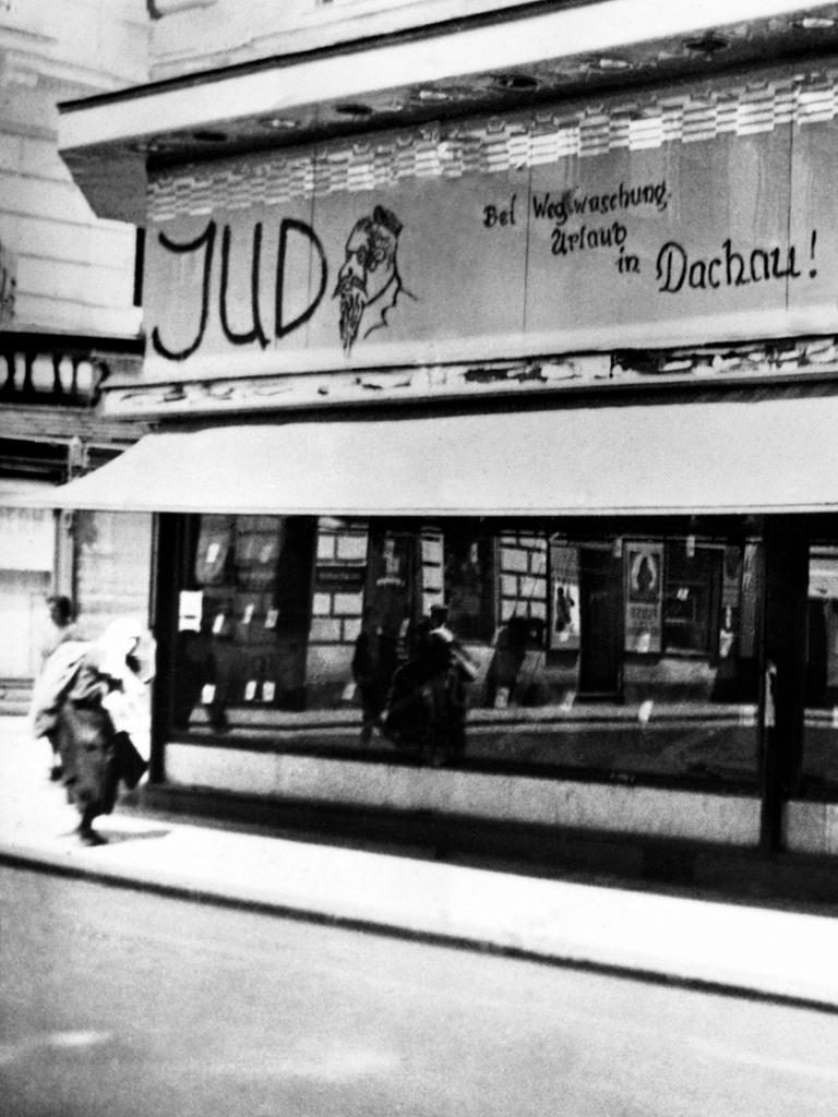 Im jüdischen Viertel in Wien, 1938. Beleidigungen und Drohungen stehen mit schwarzer Schrift über dem Schaufenster eines Geschäftes: "Bei Wegwaschung Urlaub in Dachau" und ein Männchen hängt an einem Galgen.