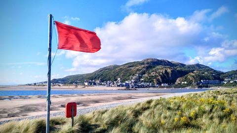 Aufnahme eines Küstenabschnitts in Wales mit Mülleimer und einer wehenden roten Flagge.