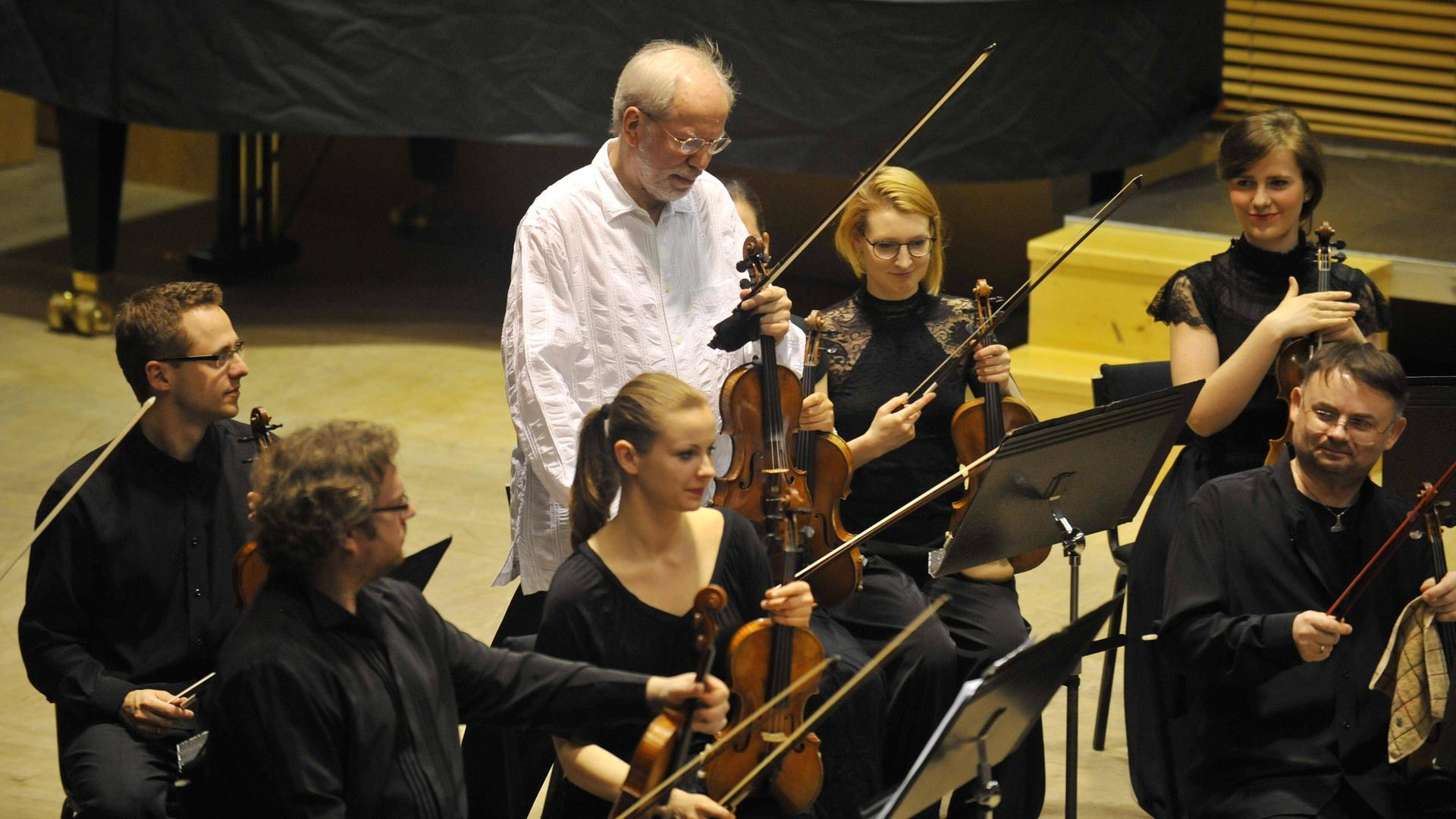Ein konzentriert auf ein Notenpult schauender weißhaariger Mann mit einer Geige in der Hand steht inmitten einer kleinen Orchestergruppe.  