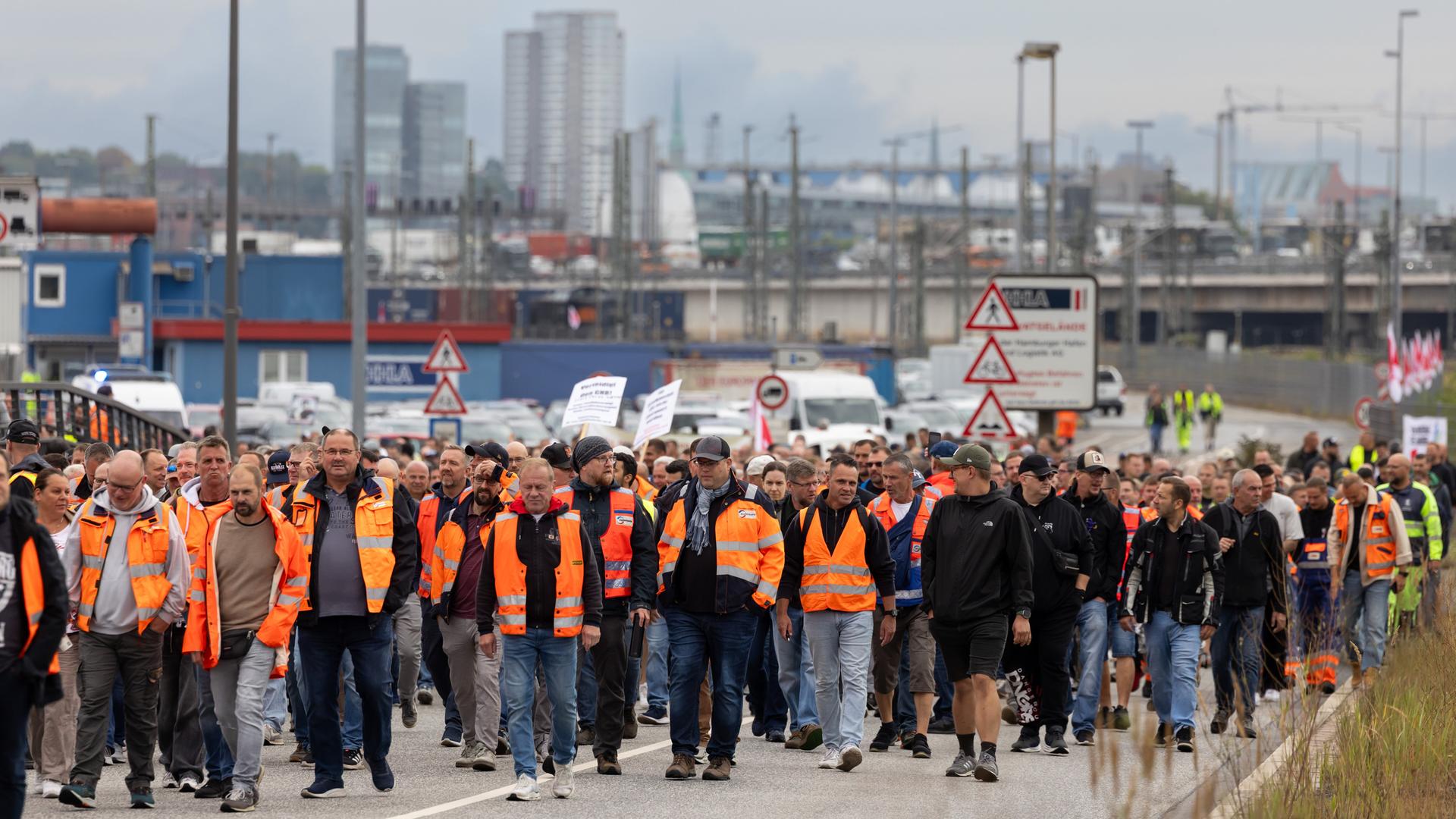 Eine große Menge an Hafenarbeitern - viele davon in orangefarbenen Warnwesten - geht einen Weg entlang. Im Hintergrund unscharf Hafenanlagen und Gebäude. Eine große Menge an Hafenarbeitern - viele davon in orangefarbenen Warnwesten - geht einen Weg entlang. Im Hintergrund unscharf Hafenanlagen und Gebäude.