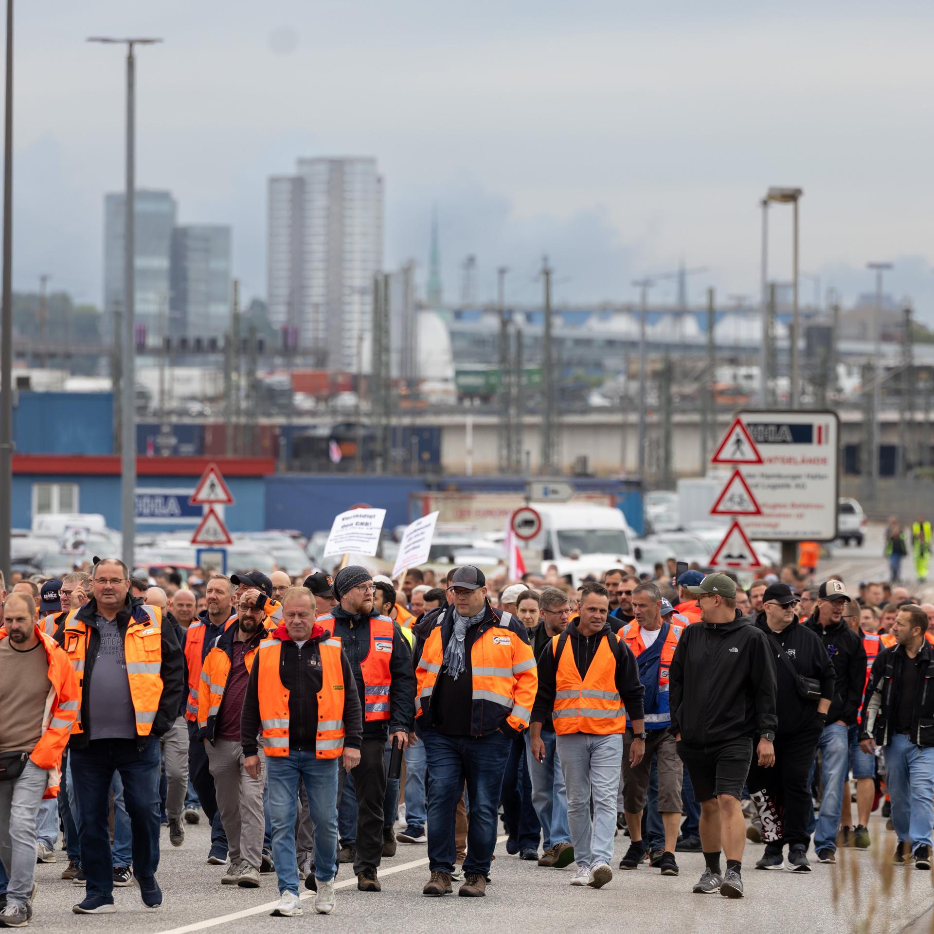 Eine große Menge an Hafenarbeitern - viele davon in orangefarbenen Warnwesten - geht einen Weg entlang. Im Hintergrund unscharf Hafenanlagen und Gebäude.