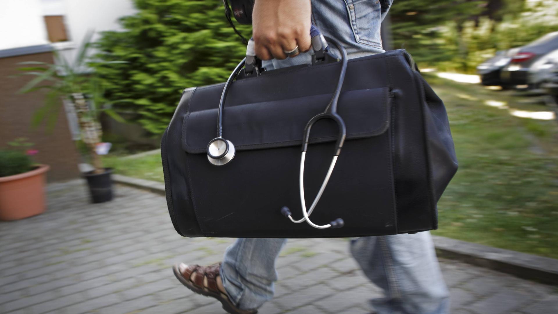 Ein Mann trägt einen schwarzen Arztkoffer, aus dem ein Stetoskop hängt eine Einfahrt entlang. 