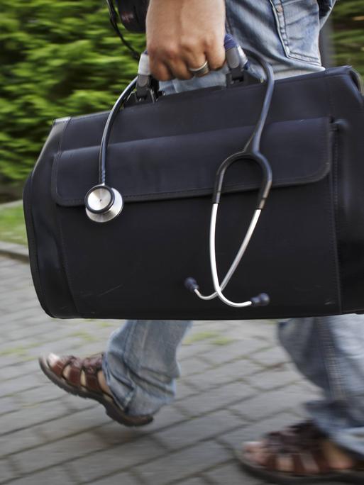 Ein Mann trägt einen schwarzen Arztkoffer, aus dem ein Stetoskop hängt eine Einfahrt entlang. 