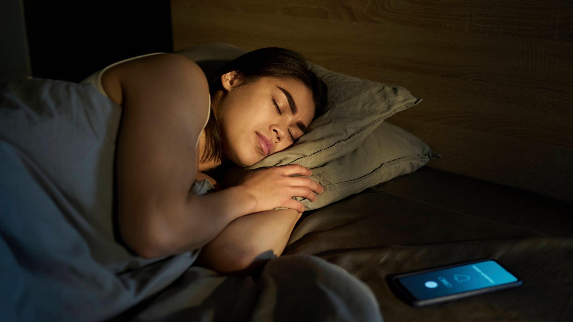 Frau schläft, während das Smartphone neben ihr im Bett Alarm schlägt.