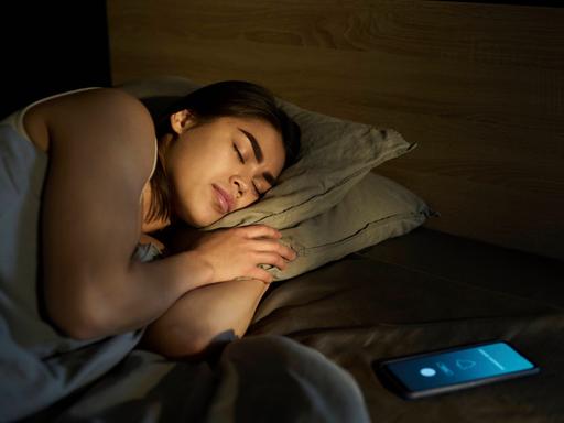 Frau schläft, während das Smartphone neben ihr im Bett Alarm schlägt.