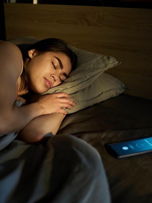 Frau schläft, während das Smartphone neben ihr im Bett Alarm schlägt.