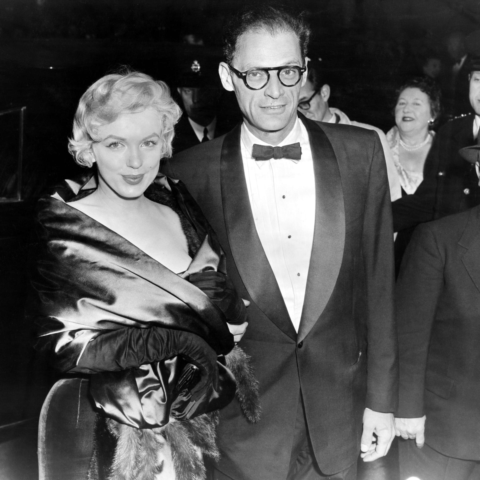 Schwarzweiß Foto von Marilyn Monroe and Arthur Miller aus dem Jahre 1956. Das Paar trägt schicke Abendgarderobe und dreht sich, vermutlich bei einem Empfang lächelnd zu der Kamera.