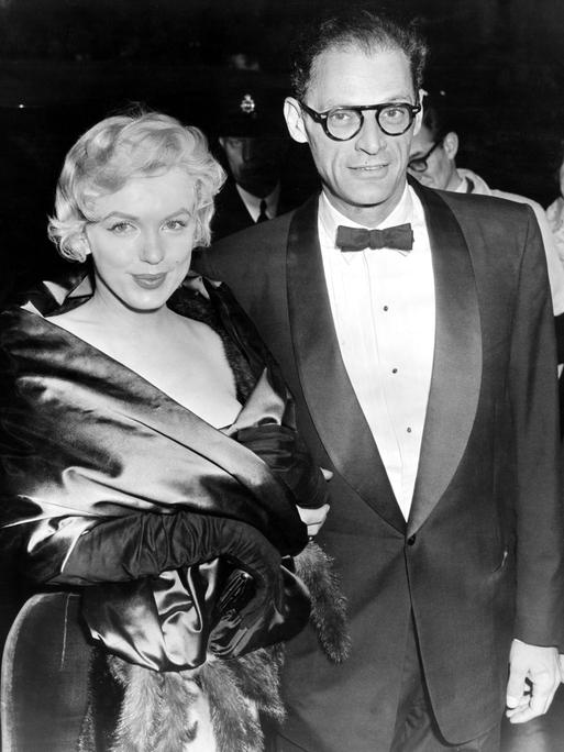 Schwarzweiß Foto von Marilyn Monroe and Arthur Miller aus dem Jahre 1956. Das Paar trägt schicke Abendgarderobe und dreht sich, vermutlich bei einem Empfang lächelnd zu der Kamera.