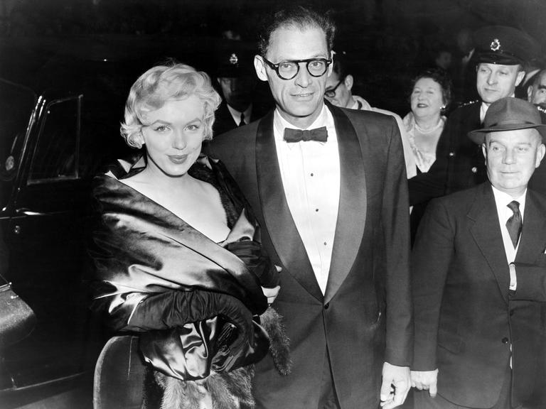 Schwarzweiß Foto von Marilyn Monroe and Arthur Miller aus dem Jahre 1956. Das Paar trägt schicke Abendgarderobe und dreht sich, vermutlich bei einem Empfang lächelnd zu der Kamera.