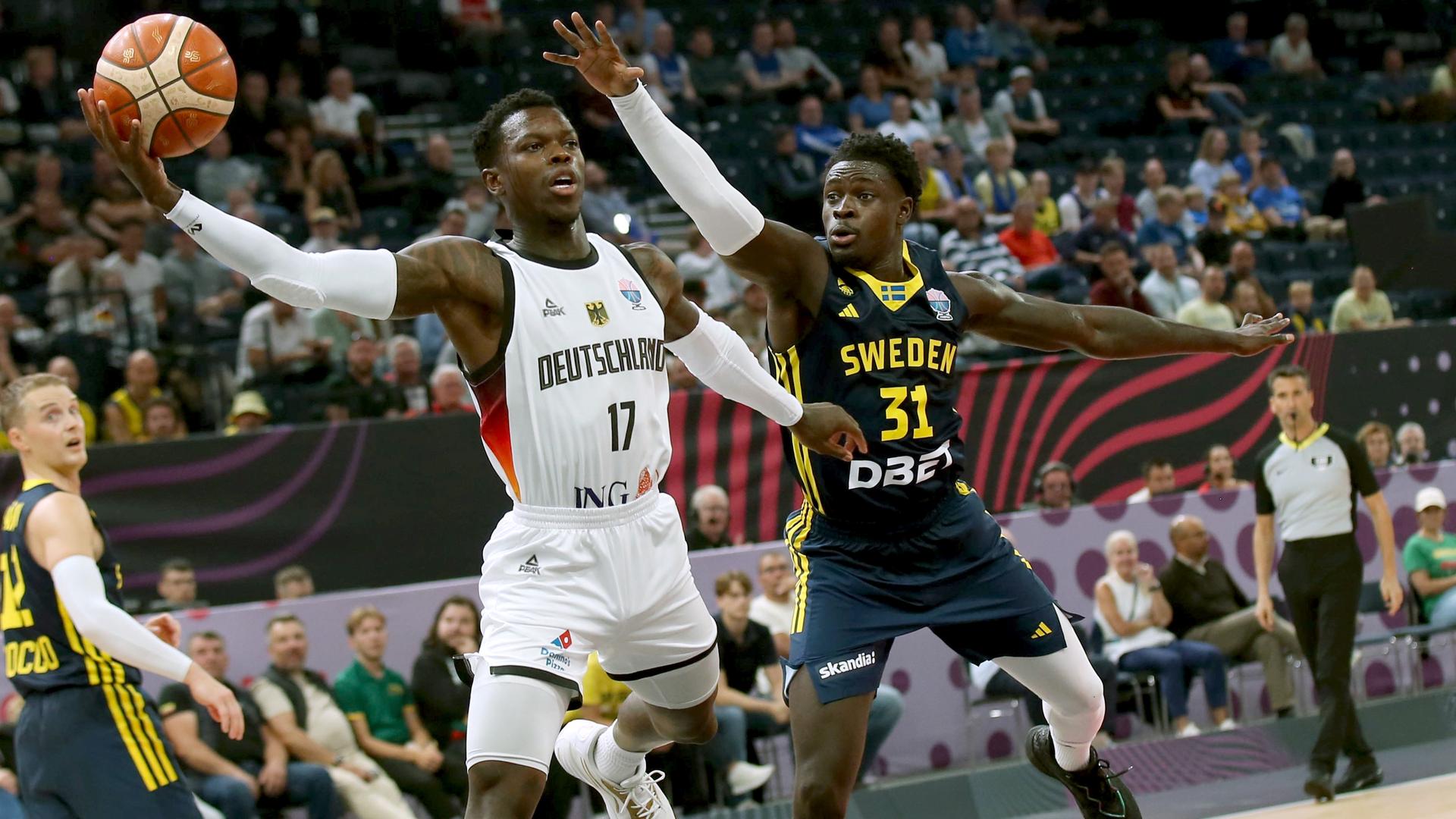 Das Bild zeigt Baskeballer Dennis Schröder beim Korbwurf, neben ihm ist ein Gegenspieler zu sehen. Das Bild zeigt Baskeballer Dennis Schröder beim Korbwurf, neben ihm ist ein Gegenspieler zu sehen.
