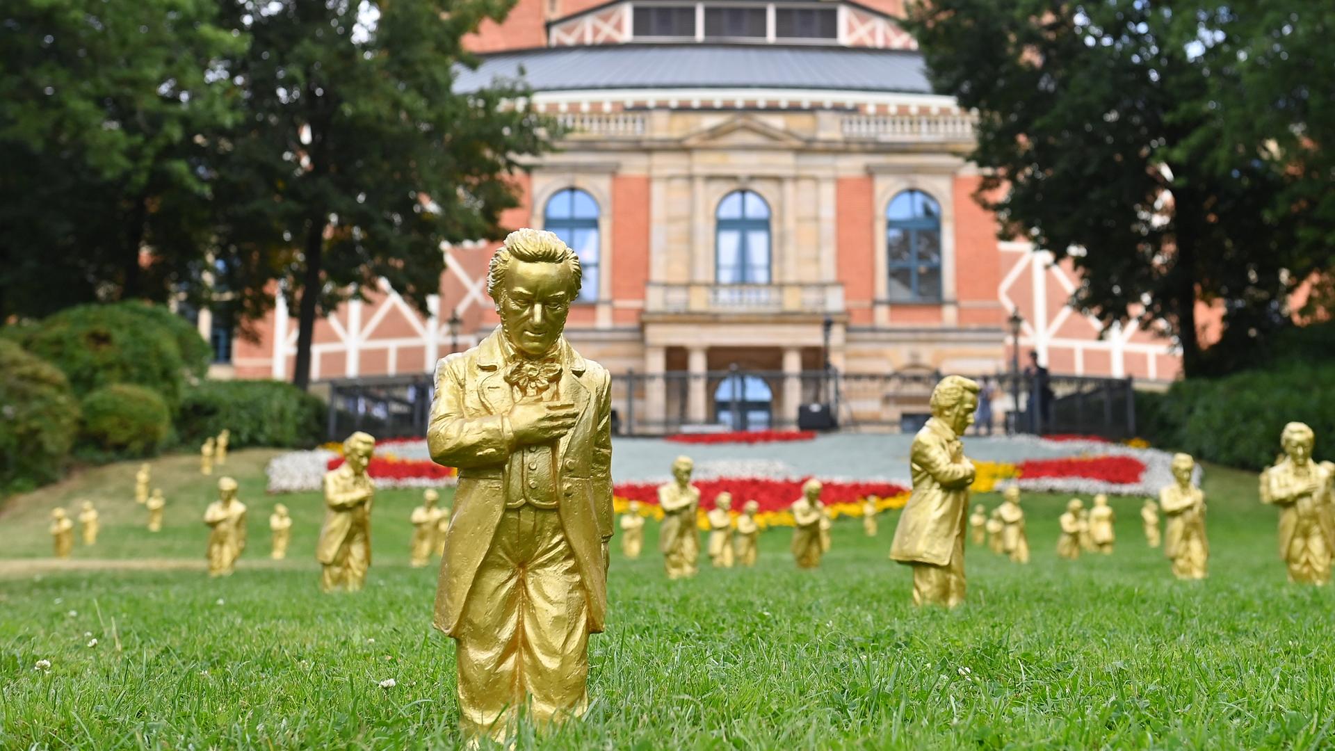 Goldene Richard-Wagner-Figuren stehen am 25.7.2024  im grünen Gras vor dem Festspielhaus in Bayreuth.
