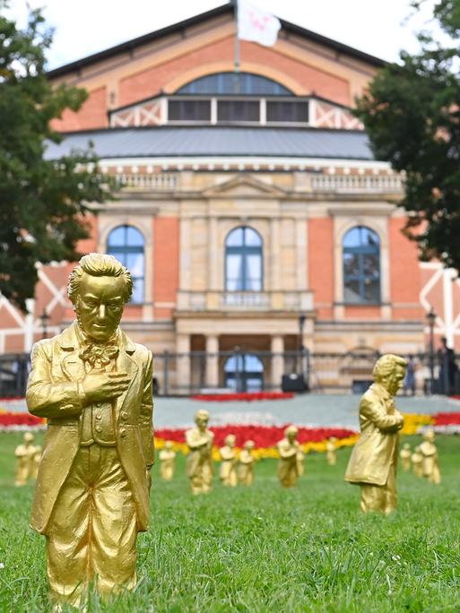 Goldene Richard-Wagner-Figuren stehen am 25.7.2024  im grünen Gras vor dem Festspielhaus in Bayreuth.
