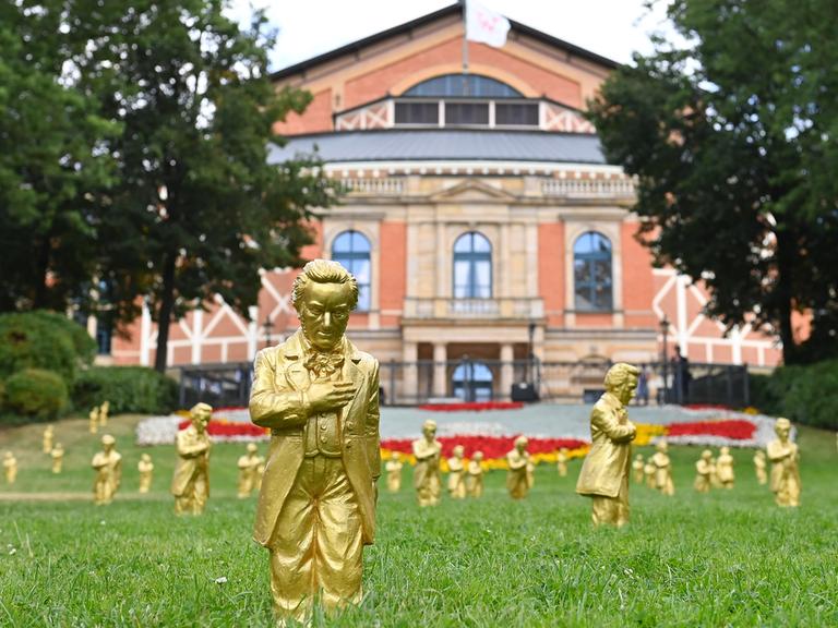 Goldene Richard-Wagner-Figuren stehen am 25.7.2024  im grünen Gras vor dem Festspielhaus in Bayreuth.
