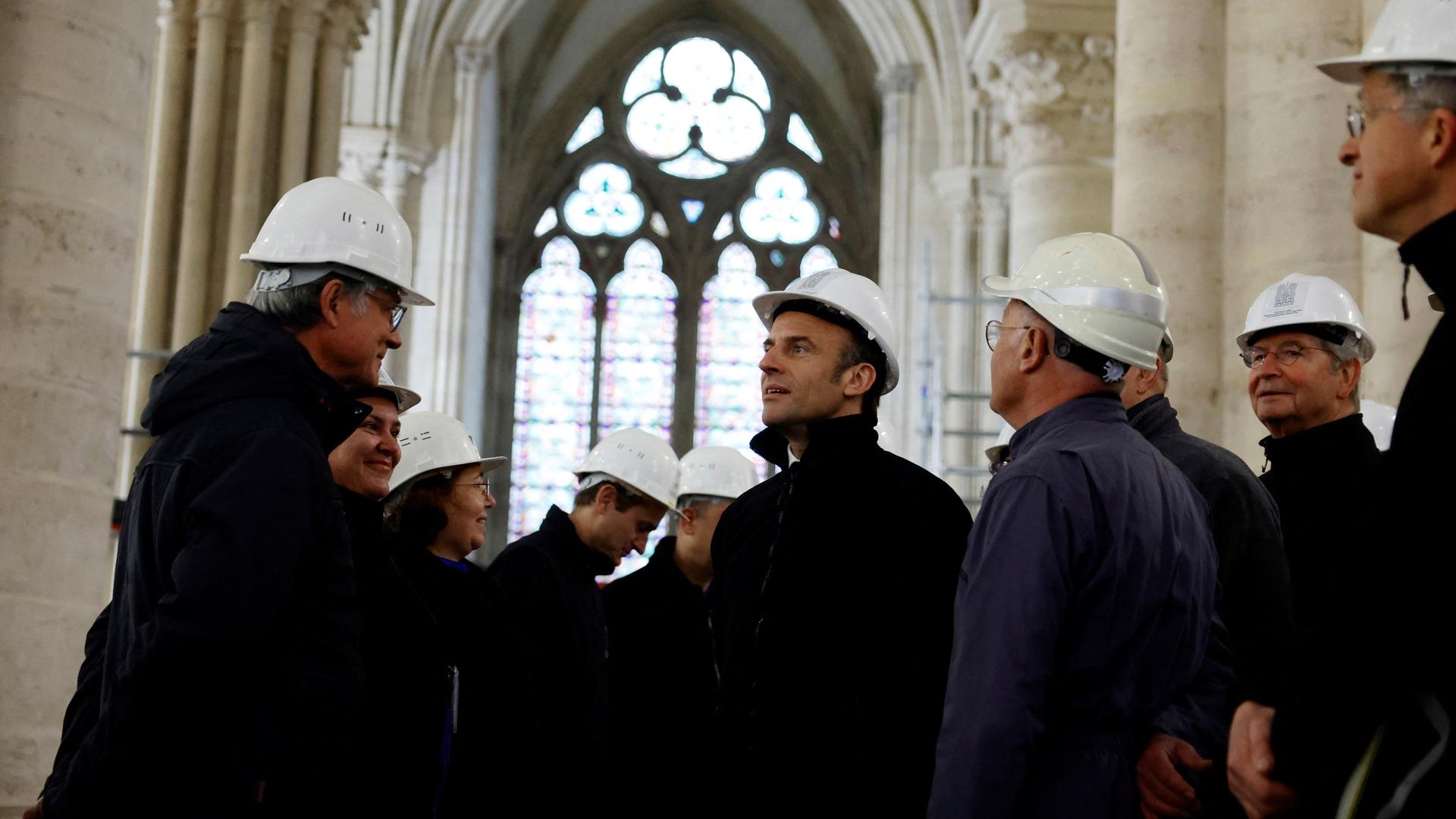 Macron und zahlreiche weitere Personen stehen mit weißen Bauhelmen in der Kathedrale. Im Hintergrund ein Kirchenfester.