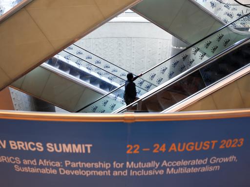 Mehrere Rolltreppen führen nach oben und unten; ein Mann steht auf einer Rolltreppe. Vorne im Foto ist ein Banner zu sehen mit der Ankündigung BRICS SUMMIT.