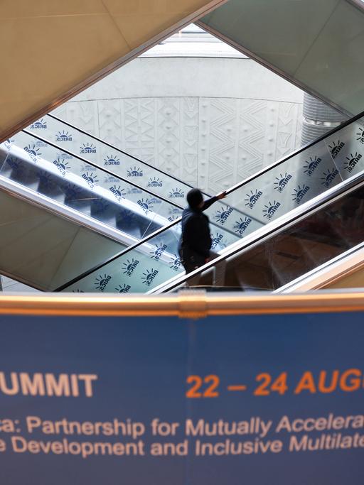 Mehrere Rolltreppen führen nach oben und unten; ein Mann steht auf einer Rolltreppe. Vorne im Foto ist ein Banner zu sehen mit der Ankündigung BRICS SUMMIT.