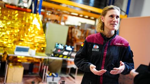 Anika Mehlis steht in einer Jacke, die ihren Namen trägt, in einem Raum und spricht gestikulierend.
