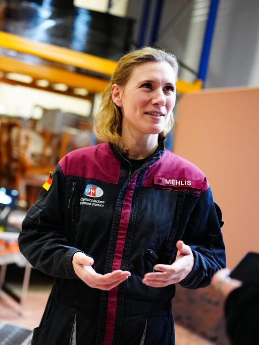 Anika Mehlis steht in einer Jacke, die ihren Namen trägt, in einem Raum und spricht gestikulierend.