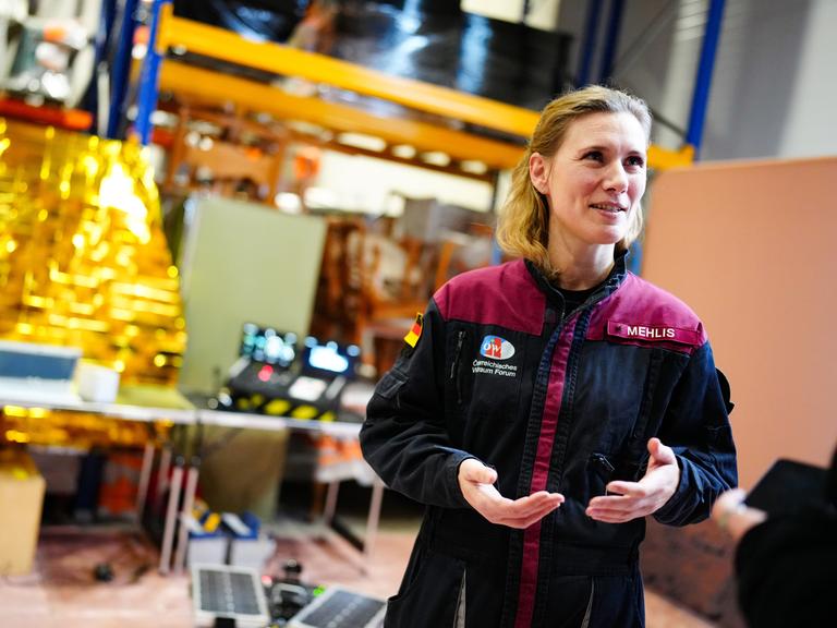 Anika Mehlis steht in einer Jacke, die ihren Namen trägt, in einem Raum und spricht gestikulierend.