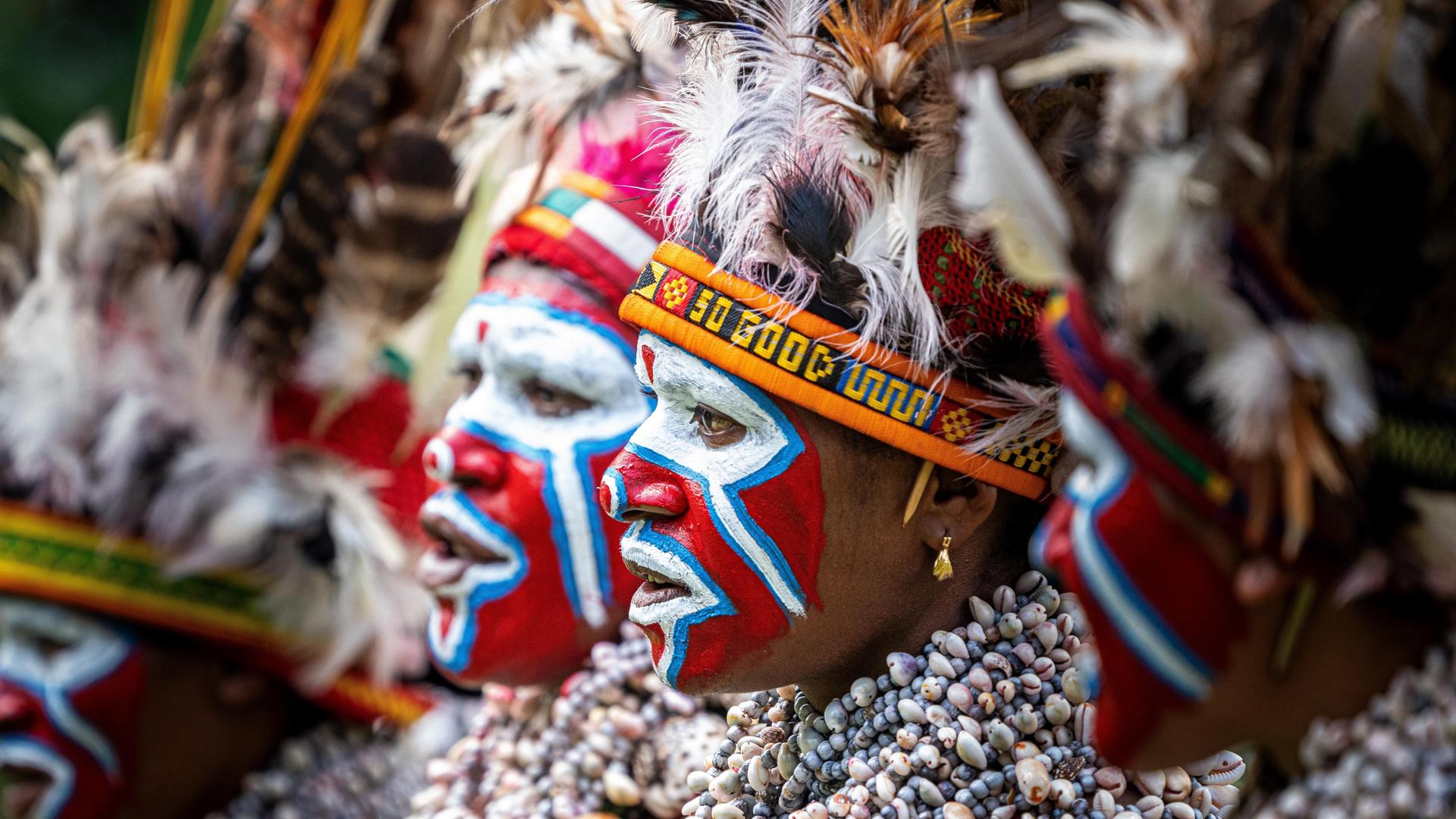 Mehrere Frauen stehen nebeneinander und tragen auffällige rote, blaue und weiße Gesichtsbemalung, hohe Kopfbedeckungen mit Federn sowie dichte Ketten aus Muscheln.