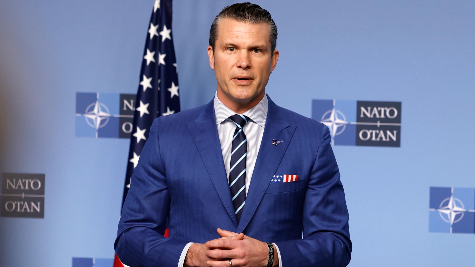 US-Verteidigungsminister Hegseth steht beim NATO-Treffen in Brüssel vor einer blauen Wand mit dem Logo der Organisation. Hinter ihm steht eine US-Fahne.