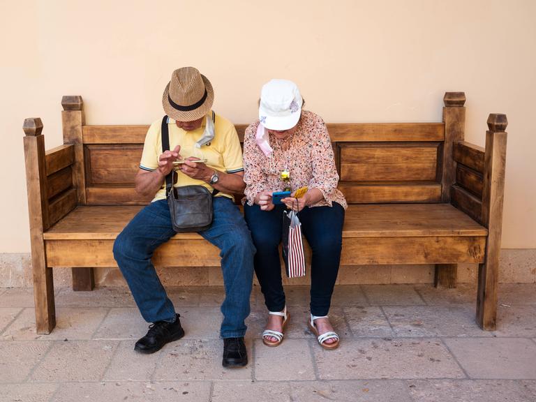 Eine ältere Frau und ein älterer Mann sitzen auf einer Holzbank und blicken auf ihre Smartphones