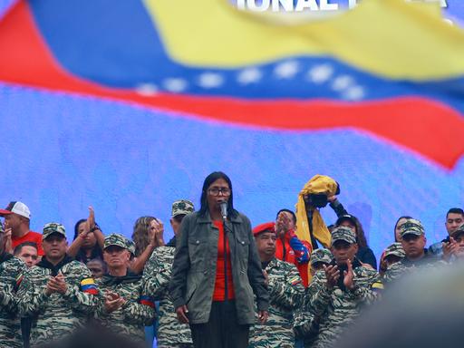 Delcy Rodriguez steht vor einer Gruppe in militärischer Kleidung. Über ihnen weht die venezolanische Flagge.  