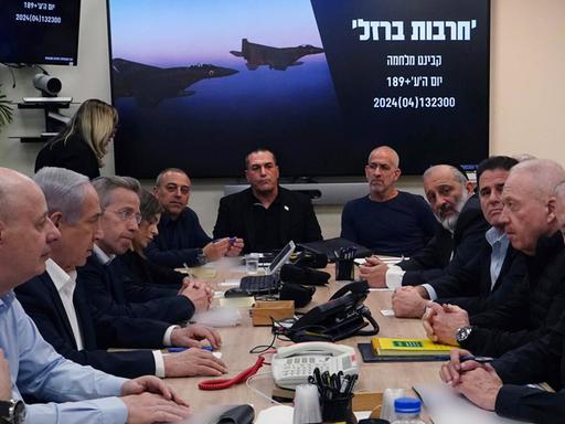 Israels Ministerpräsident Netanjahu sitzt mit seinem Kriegskabinett, das aus mehrere Personen besteht, zu Beratungen um einen Tisch. Im Hintergrund ist auf einem Display an der Wand ein hebräischer Schriftzug und ein Kampfbomber zu sehen. 