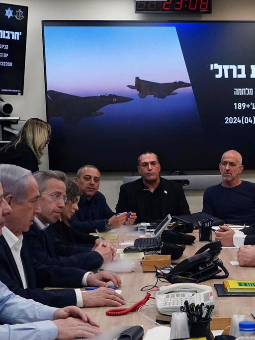 Israels Ministerpräsident Netanjahu sitzt mit seinem Kriegskabinett, das aus mehrere Personen besteht, zu Beratungen um einen Tisch. Im Hintergrund ist auf einem Display an der Wand ein hebräischer Schriftzug und ein Kampfbomber zu sehen. 