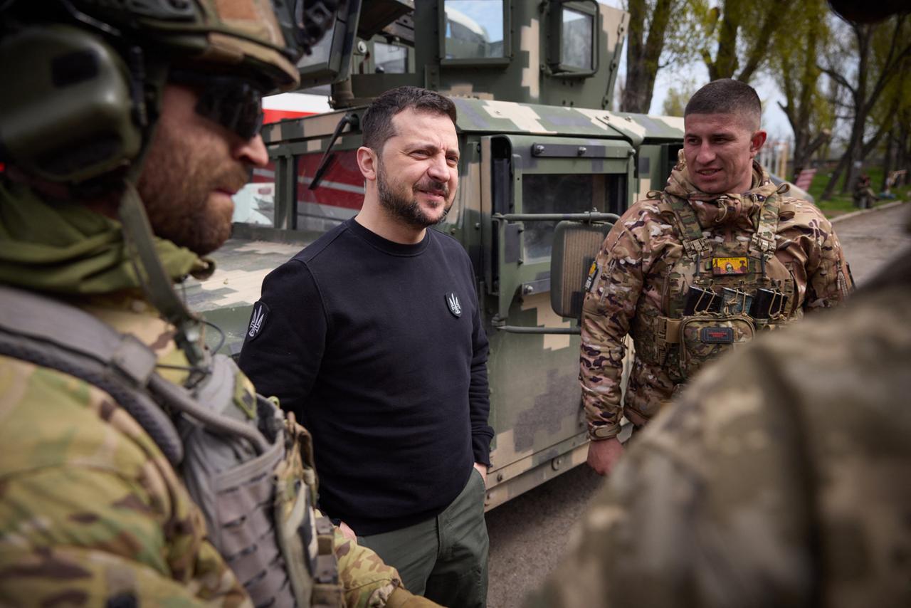 Präsident Selenskyj steht mit Soldaten zusammen und unterhält sich. Er hat einen schwarzen Pullover an. 