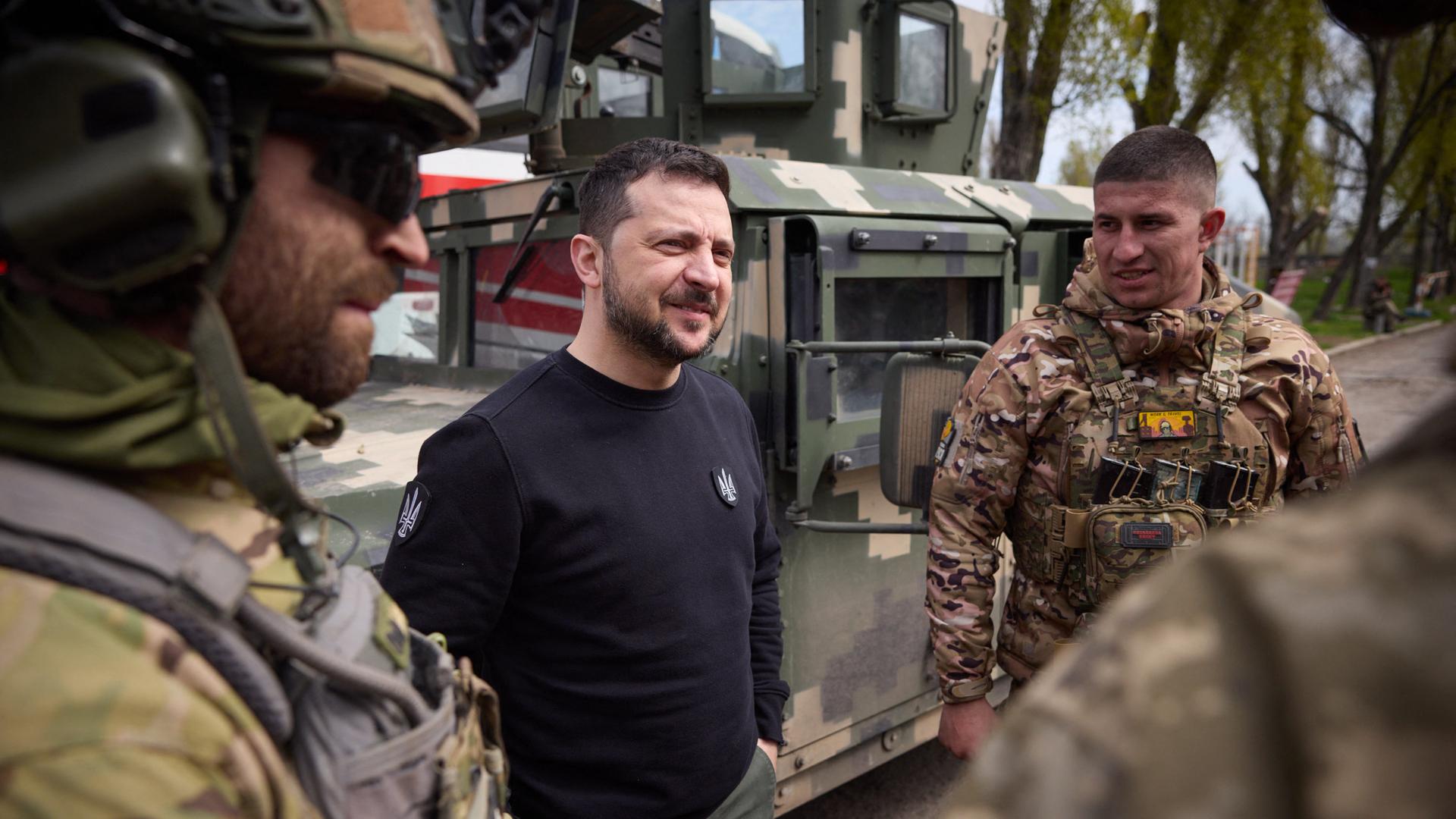 Präsident Selenskyj steht mit Soldaten zusammen und unterhält sich. Er hat einen schwarzen Pullover an.