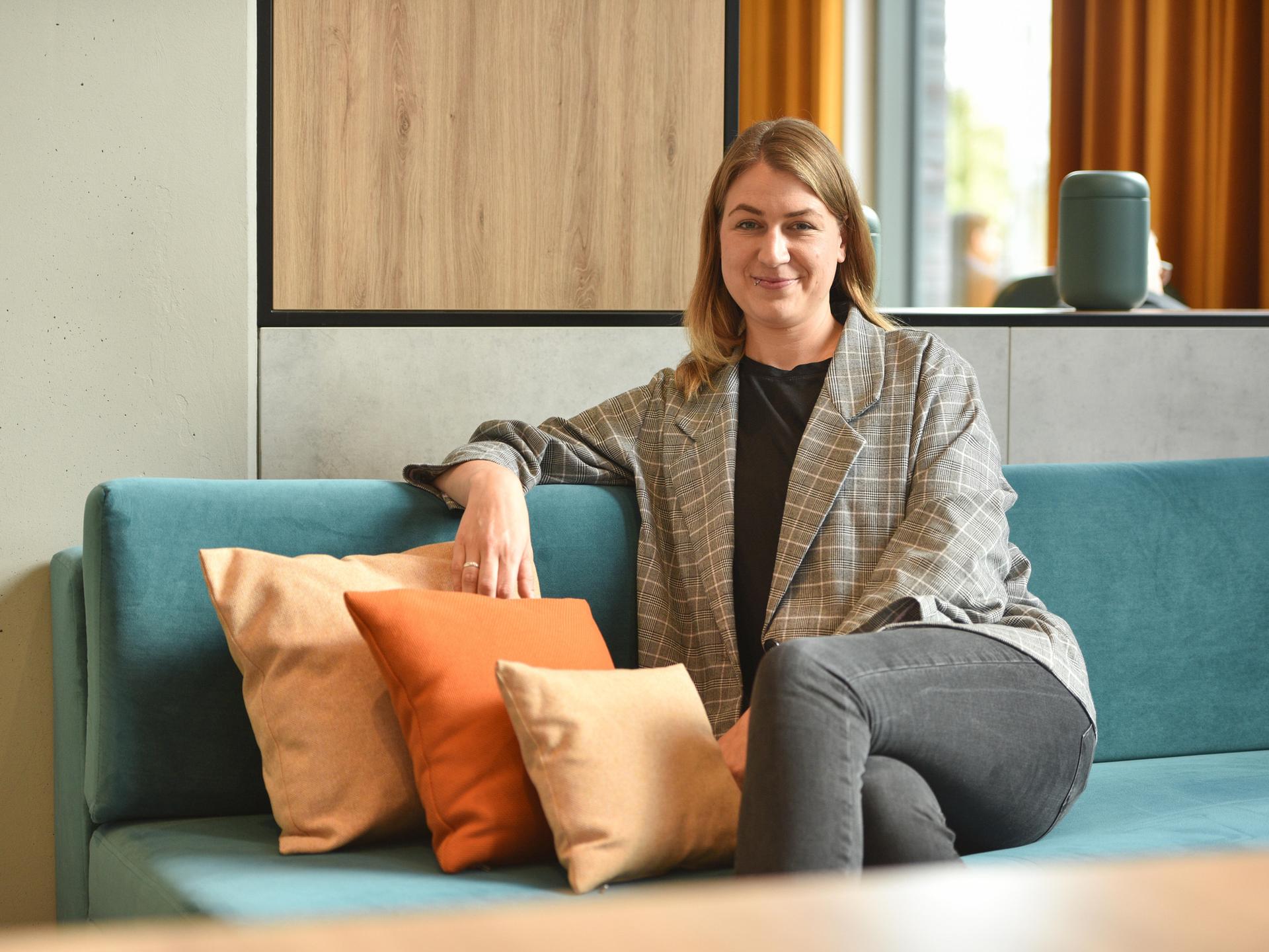 Ina Jäckel sitzt auf einem Sofa und schaut in die Kamera. Sie hat dunkelblondes Haar und trägt einen karierten Blazer.