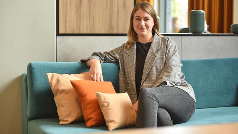 Ina Jäckel sitzt auf einem Sofa und schaut in die Kamera. Sie hat dunkelblondes Haar und trägt einen karierten Blazer.