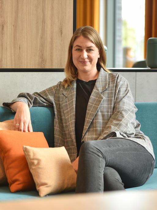 Ina Jäckel sitzt auf einem Sofa und schaut in die Kamera. Sie hat dunkelblondes Haar und trägt einen karierten Blazer.