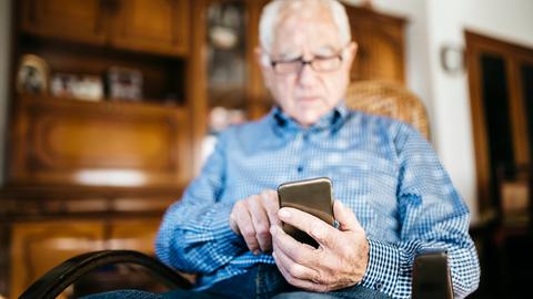 Ein älterer Mann sitzt in einem Stuhl und schaut hinab auf sein Smartphone. Ein älterer Mann sitzt in einem Stuhl und schaut hinab auf sein Smartphone.