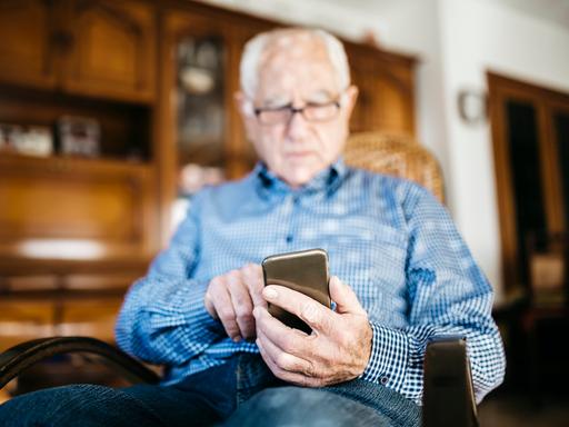 Ein älterer Mann sitzt in einem Stuhl und schaut hinab auf sein Smartphone.