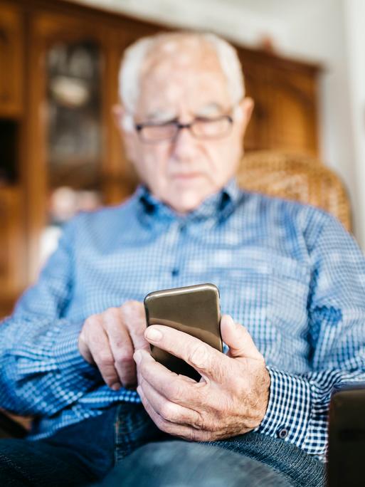 Ein älterer Mann sitzt in einem Stuhl und schaut hinab auf sein Smartphone.