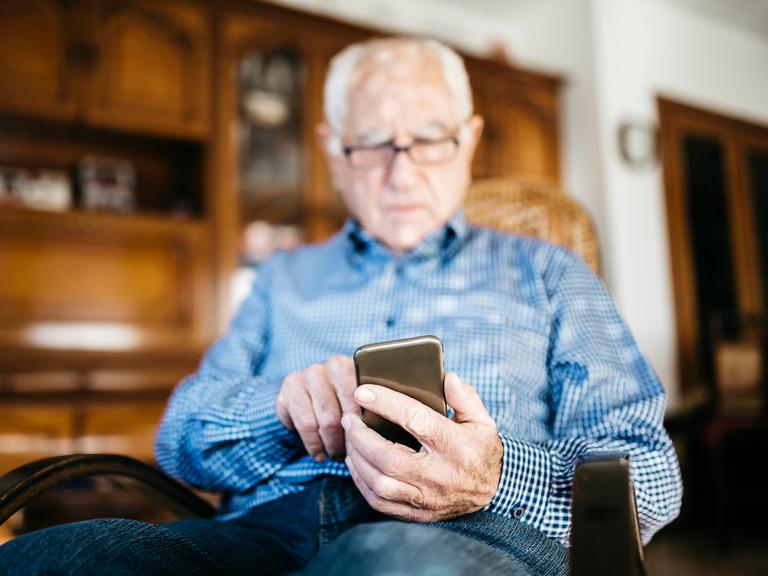 Ein älterer Mann sitzt in einem Stuhl und schaut hinab auf sein Smartphone.