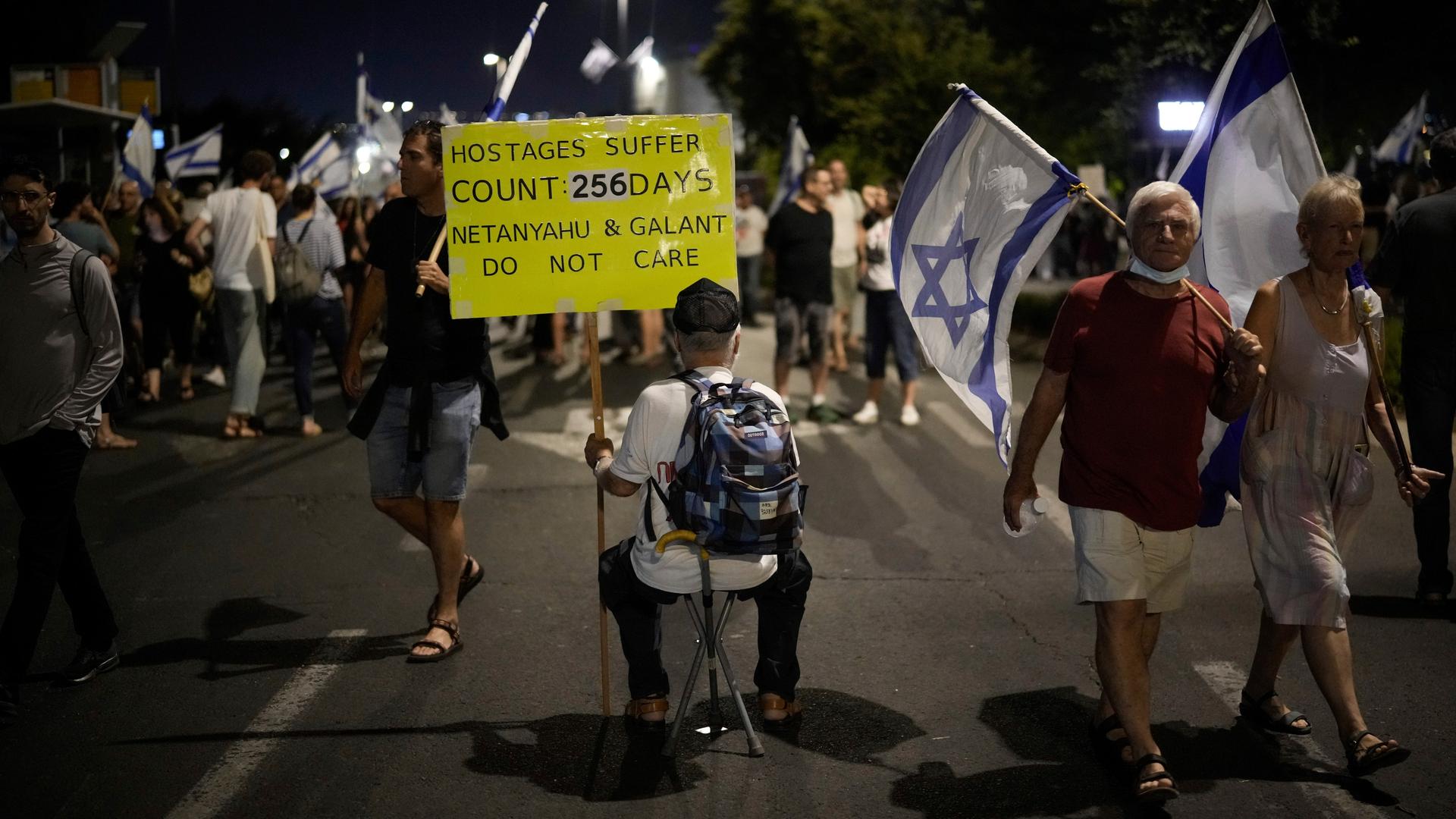 Ein Mann sitzt mit einem Transparent auf der Straße; um ihn herum sind Menschen mit israelischen Flaggen unterwegs.