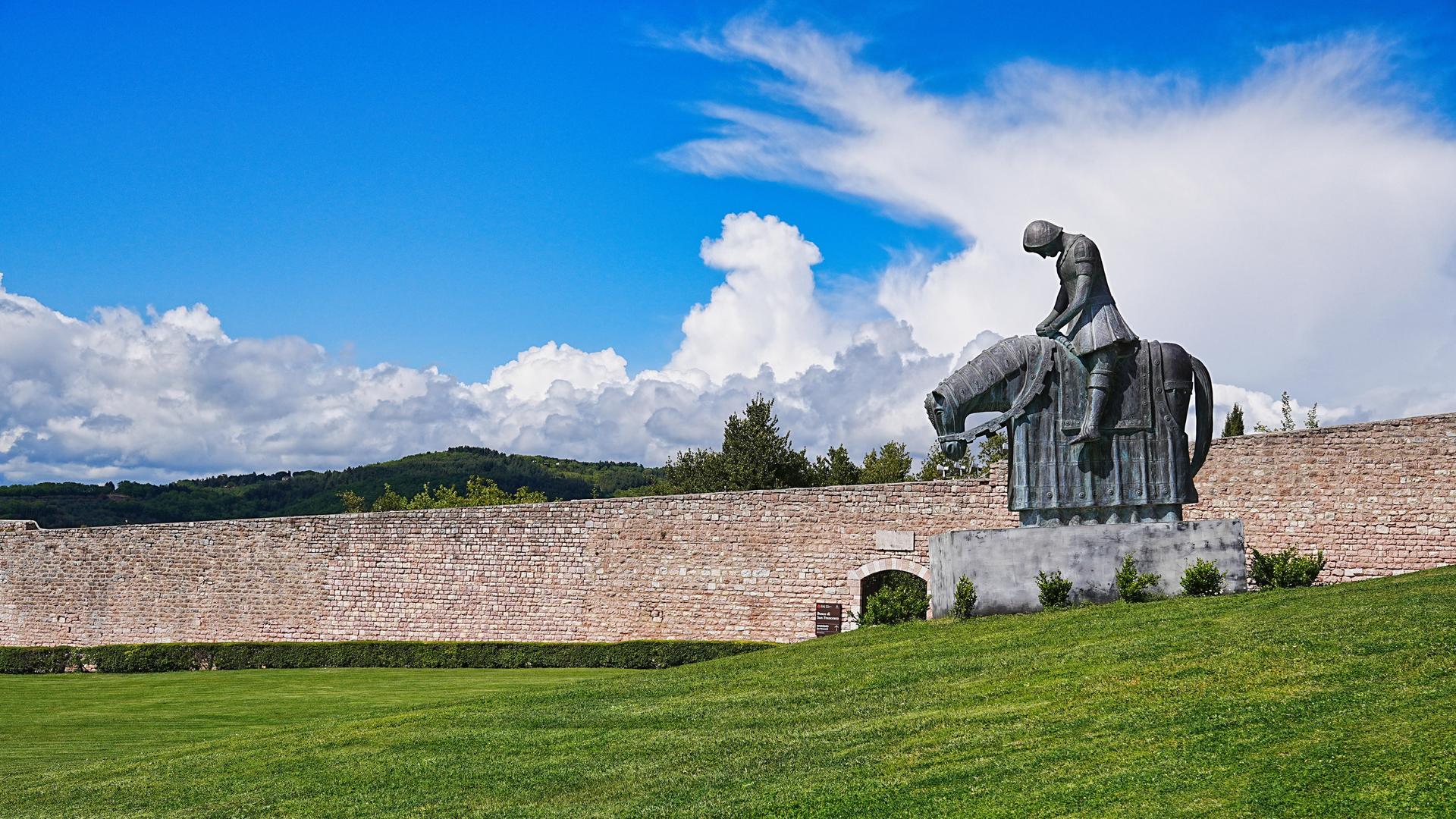 Franziskusstatue von Norberto Proietti: Franziskus reitet in voller Rüstung, den Oberkörper vornüber gebeugt. Er kehrt krank und demütig aus dem Krieg nach Assisi zurück.