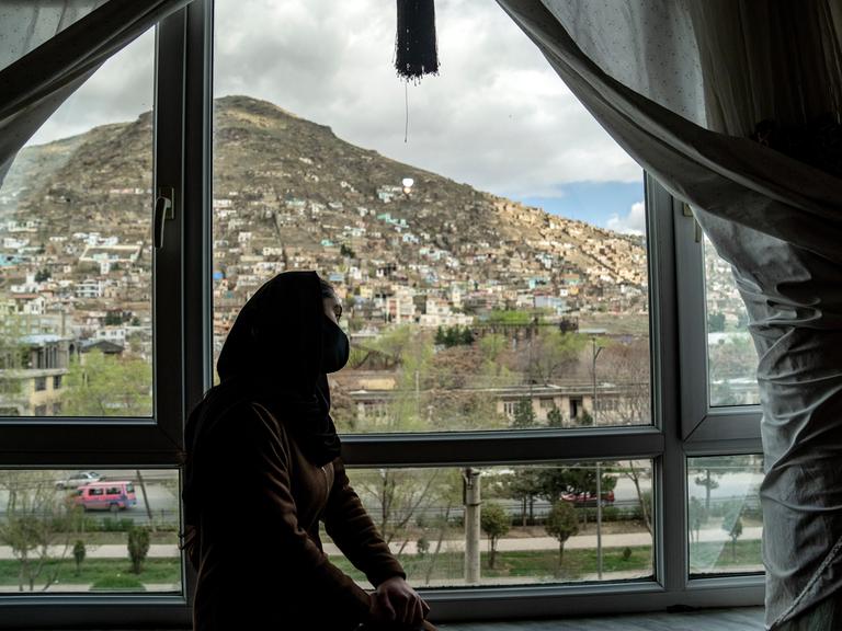 In Kabul sitzt ein junges Mädchen verschleiert an einem Fenster und blickt auf die Stadt.