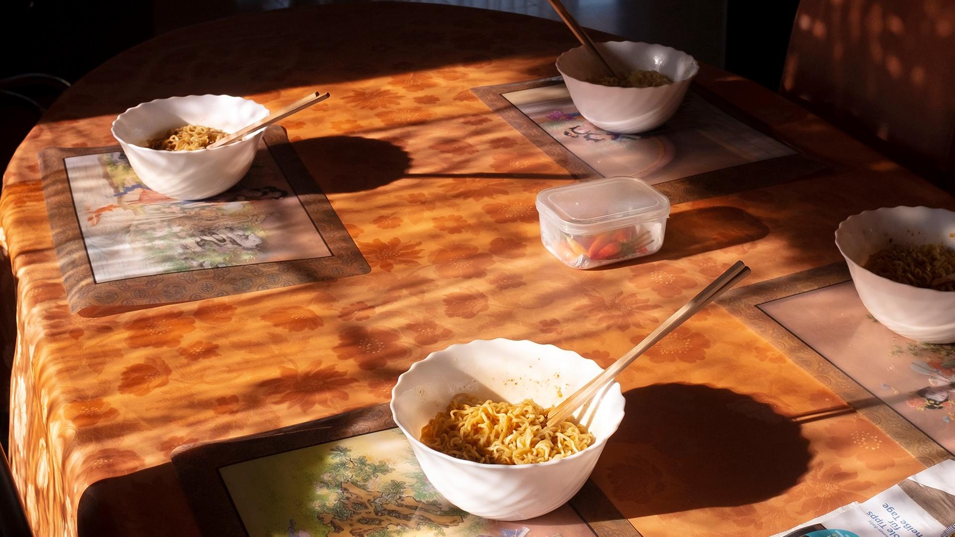 Eine Fotografie von Mika Sperling mit dem Titel "Lunch" zeigt in einer Wohnung einen Esstisch aus hellem Holz, auf dem zwei Schalen mit asiatischen Nudeln angerichtet sind.