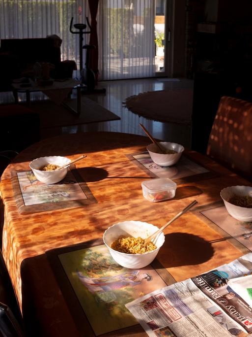 Eine Fotografie von Mika Sperling mit dem Titel "Lunch" zeigt in einer Wohnung einen Esstisch aus hellem Holz, auf dem zwei Schalen mit asiatischen Nudeln angerichtet sind.