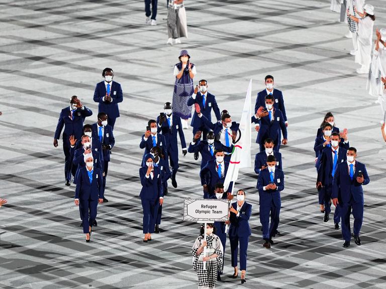 Zu sehen ist die Eröffnungsfeier bei den Olympischen Spielen in Tokio 2021, als gerade das damalige Refugee Olympic Team einläuft und dabei die Olympische Fahne hochhält.